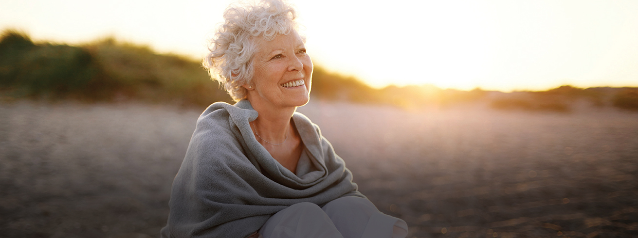 Female senior sitting on the beach smiling looking ahead in the distance with the sun in the horizon